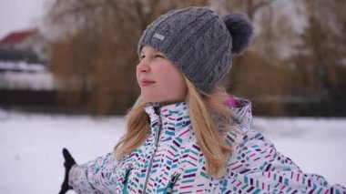 Satisfied teenage pretty girl stretching hands looking away smiling standing in winter park on cloudy day. Portrait of happy carefree relaxed Caucasian teenager enjoying weekend outdoors. Slow motion