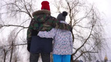 Back view teenage boy hugging girl in slow motion standing outdoors. Confident Caucasian teen couple dating in cloudy park looking away embracing