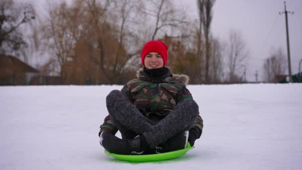 Портрет Смеющегося Расслабленного Подростка Сидящего Зеленых Санях Белом Снегу Смотрящего — стоковое видео