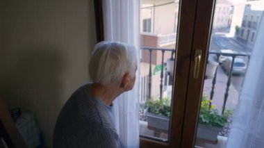 Old sad Caucasian grey-haired woman looking out window looking at camera turning. Portrait of senior wrinkled retiree thinking and posing indoors at home on sunny summer day