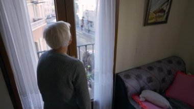 High angle view old grey-haired Caucasian woman standing up from chair opening balcony door. Senior female retiree enjoying summer day at home indoors. Leisure and retirement