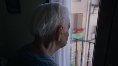 Close-up grey-haired wrinkled old woman standing in darkness looking out window. Unrecognizable Caucasian female senior retiree admiring sunny summer street standing at home indoors