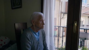 Sad thoughtful wrinkled old woman sitting on chair at balcony door looking around and looking out the window. Portrait of senior Caucasian female retiree thinking indoors at home