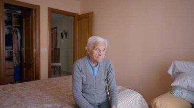 Portrait of thoughtful old woman looking away and looking at camera sitting on bed indoors. Medium shot of female Caucasian senior retiree posing at home