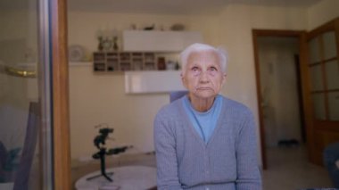 Front view portrait of grey-haired wrinkled old woman looking up and looking at camera sitting indoors. medium shot of confident Caucasian female retiree posing at home. Relaxation and lifestyle