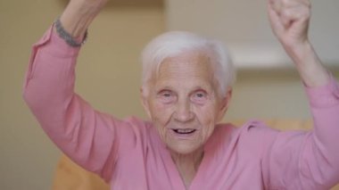 Portrait of grey-haired happy old woman with grey eyes looking at camera rejoicing watching match on TV. Cheerful positive Caucasian retiree having fun enjoying leisure at home indoors posing