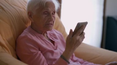 Side angle view portrait of confident old woman talking looking at smartphone app sitting on armchair. Relaxed Caucasian senior female retiree using modern technologies at home indoors