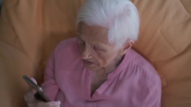 High angle view portrait of wrinkled old Caucasian woman talking using video chat in smartphone app. Senior grey-haired retiree chatting online sitting indoors at home. Convenience and aging concept