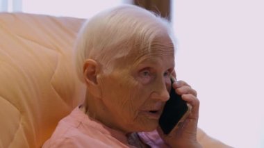 Close-up old Caucasian woman talking on phone sitting indoors at home. Serious senior grey-haired retiree chatting using smartphone. Lifestyle and modern technologies