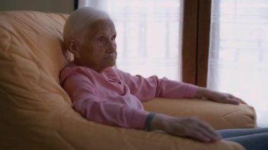 Relaxed retiree tapping hand on armrest sitting in armchair indoors. Side view portrait of thoughtful Caucasian old woman enjoying leisure at home indoors