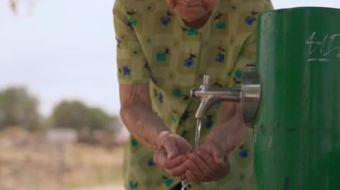Senior female retiree scoop up water in the palm drinking standing at faucet outdoors. Front view old Caucasian wrinkled woman enjoying refreshing liquid on summer day