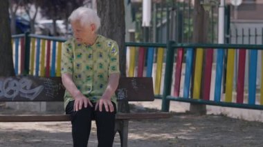 Portrait of old woman sitting on bench with colorful fence at background. Thoughtful relaxed Caucasian retiree enjoying summer vacations resting outdoors looking around