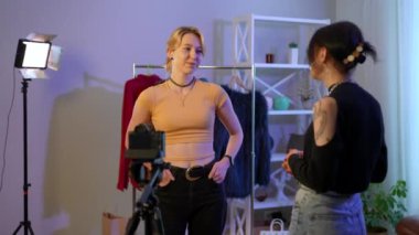 Medium shot portrait of smiling slim young Caucasian woman talking with lady standing at rack with clothes in studio. Happy confident blogger discussing fashion with guest indoors