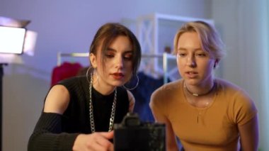 Two beautiful positive young Caucasian women talking adjusting video camera standing indoors. Confident charming blond and brunette bloggers getting ready for live streaming