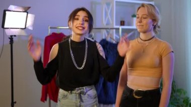 Two confident charming women talking looking at camera talking about fashion and style standing indoors. Medium shot of confident beautiful Caucasian bloggers posing filming vlog at rack with clothing