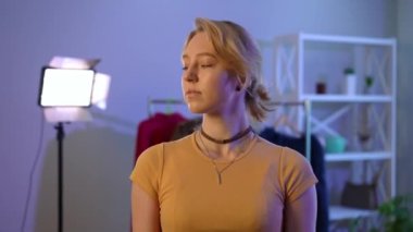 Attractive coquette young slim woman looking away and looking at camera standing indoors. Zoom in portrait of beautiful blond Caucasian lady with green eyes posing at light stand. Blogging femininity