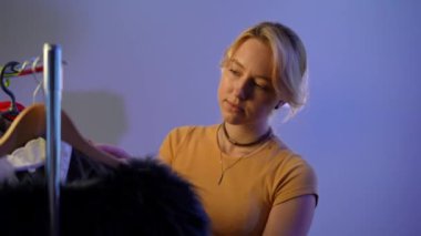 Young charming Caucasian blond woman choosing dress in the morning at home indoors. Portrait of thoughtful focused attractive millennial lady thinking standing at rack with clothes hangers