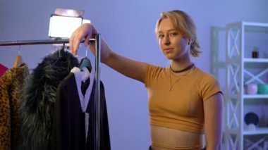 Medium shot portrait of satisfied young slim beautiful woman standing at rack with clothes looking at camera. Happy confident Caucasian lady posing indoors with fashionable outfits. Slow motion