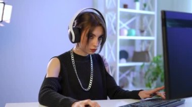 Zoom in to serious female gamer in headphones sitting at PC playing video game and looking at camera. Absorbed Caucasian young gorgeous woman gaming online in living room at home posing