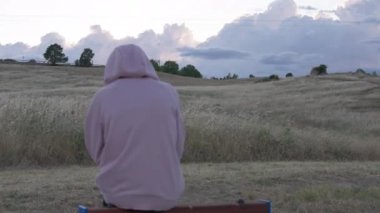 Back view lonely old woman sitting on overcast windy day outdoors on meadow with hills thinking. Senior Caucasian female retiree on the left resting on bench in field in Spain. Tourism and loneliness