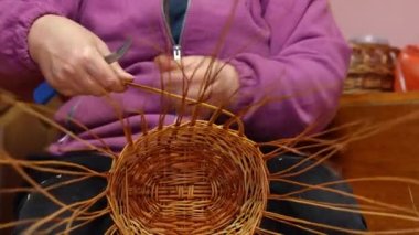Hands of plus-size Caucasian woman bending strands making wicker basket. Unrecognizable artisan creating design of handmade decoration sitting indoors