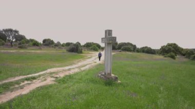 Wide shot summer spring lavender field with big cross and unrecognizable old woman walking away on countryside road. Religious symbol on picturesque landscape