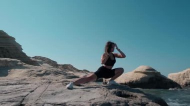 Wide shot portrait of fit slim sportswoman stretching in side lunges outdoors. Front view of brunette beautiful Caucasian woman working out at Mediterranean sea on rocks at background of blue sky