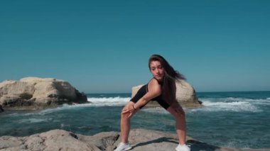 Portrait of gorgeous fit sportswoman looking at camera working out at background of Mediterranean sea waves splashing on rocks. Wide shot front view of slim motivated Caucasian woman posing training