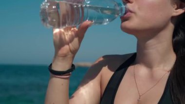 Close-up unrecognizable tanned young woman drinking refreshing water at background of blue Mediterranean sea waves and cloudless summer sky. Relaxed confident lady enjoying drink in sunshine
