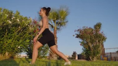 Side view wide shot of fit sportswoman exercising in lunges outdoors in sunshine. Motivated slim beautiful Caucasian lady training at sunset on Cyprus