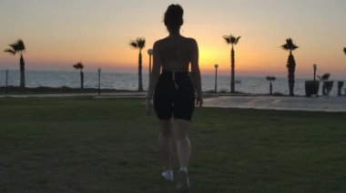 Tracking shot of confident young woman walking to golden sunset on Mediterranean sea. Back view slim Caucasian tourist strolling outdoors in twilight. Tourism and lifestyle concept