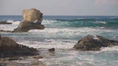 Turkuaz mavi dalgalar Akdeniz açıklarında kayaların üzerinde yuvarlanıyor. Bulutlu bir günde köpüklü okyanus suyu sıçrıyor