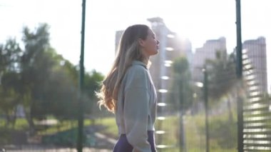 Güneş ışınlarıyla yürüyen kendine güvenen bir sporcunun çitleri aşarken yan görüntüsü. Güzel, beyaz, genç bir kadın idmandan sonra dışarıda geziniyor. Yaşam tarzı ve motivasyon kavramı
