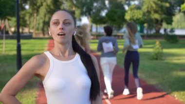 Solda duran gülümseyen genç kadın sporcu kadınlar arka planda maraton koşmaya başlarken. Kendine güveni tam beyaz bir kadının portresi başka tarafa bakıyor.