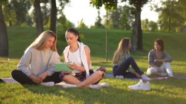 Günbatımında yeşil çimlerde oturup ağır çekimde konuşan dijital tabletli beyaz kadınlar. Bulanık arkadaşlar arka planda gün ışığında sohbet ediyorlar. Spor ve arkadaşlık kavramı