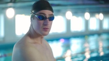 Havuz kenarında yüzme şapkalı ve gözlüklü beyaz bir adam havuz kenarında kameraya bakıp gülümsüyor. İçeride poz veren kendinden emin bir sporcunun portresi.