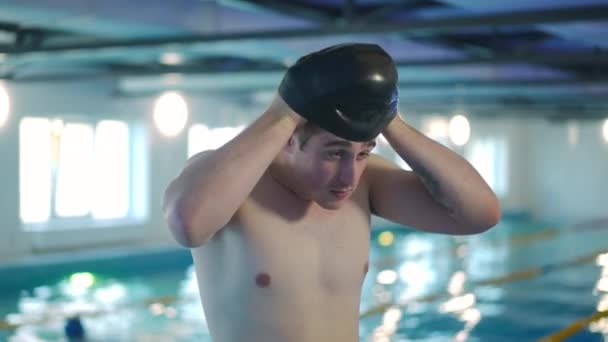 若い男は屋内プールに立って水泳キャップを置く 男性白人の自信に満ちた水泳選手の肖像訓練の準備をして取得します スポーツと健康的なライフスタイルのコンセプト — ストック動画