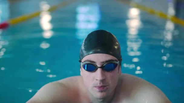 一个自信而严肃的年轻人 戴着泳镜 头戴水帽 站在室内的前视图画像 被激励的运动员看着镜头 慢动作地在游泳池里摆姿势 — 图库视频影像