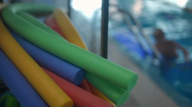 Havuz kenarında renkli yüzme çubukları var. Arka planda bulanık çocuklar suda eğitim görüyor. Yüzme havuzunda yüzülebilir oyuncaklar. Yavaş çekim