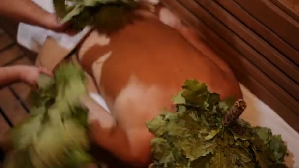 俯瞰的男人躺在俄罗斯澡堂里 按摩师用桦树扫帚给他按摩 高角镜白种人客户享受在芬兰桑拿室内的服务 — 图库视频影像