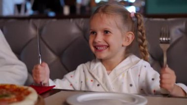 Gülümseyen neşeli bir kızın portresi çatal ve bıçakla kafeteryada tanınmayan anne babasıyla otururken görülüyor. Mutlu gülümseyen beyaz kız hafta sonu ailesiyle eğleniyor.