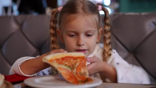 近くで見る幸せな白人の女の子おいしいピザスライス笑みを浮かべて噛むピグテールを持つ 屋内レストランでおいしい料理を楽しむ満足かわいい子供の肖像画 — ストック動画