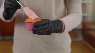 Genç bayan aşçı keki hamur fırçası kullanarak sıvıyla kaplıyor. Ön manzara tanınmaz halde, eldivenli beyaz kadın evde lezzetli fırınlar pişiriyor.