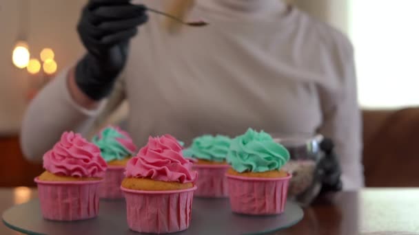 特写松饼与皇家糖衣托盘与女性的手撒播可食珍珠 难以辨认的白人妇女在室内用五颜六色的糖果球装饰烤美味的糕点 — 图库视频影像
