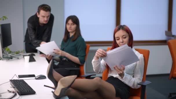 一个粗心大意的年轻女人 脚踏在桌子上到处乱扔文件 而忙碌的同事们却在背后喋喋不休 与同事一起在任的无忧无虑的白人女性雇员的画像 — 图库视频影像
