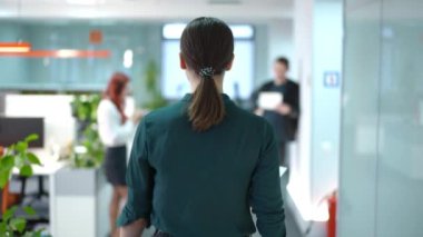 Ofise gelen kendine güvenen genç bir kadının iş yerinden ayrılan meslektaşlarını selamlarken görüntüsü. Kafkasyalı kadın çalışanın arkadan görünüşü sabahları kapalı. Yavaş çekim