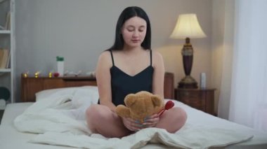 Geniş açılı, pijamalı genç bir kadın yatakta oturmuş oyuncak ayıyı kucaklıyor ve başka tarafa bakıyor. Kafkasyalı kadının portresi sabah yatak odasında oyuncakla