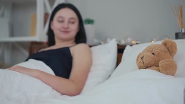 Zbliżenie Brązowy Pluszowy Miś Leżący Łóżku Zamazaną Młodą Kobietą Uśmiechniętą — Wideo stockowe