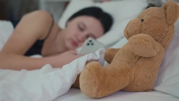 Zbliżenie Pluszowy Miś Przytulnym Łóżku Zamazaną Młodą Kobietą Przewijanie Social — Wideo stockowe