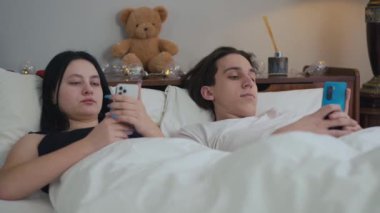 Akıllı telefon uygulaması ile sosyal medyayı kaydıran dikkatsiz, beyaz bir çift evde yatakta yatıyor. Sabahları yatak odasında sıkılmış sevgili.